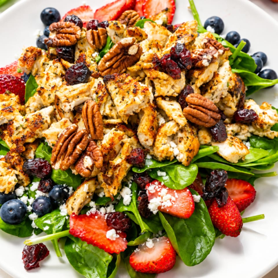 Grilled chicken, pecans, dried cranberries, berries, strawberries, and feta atop fresh spinach on a white plate.